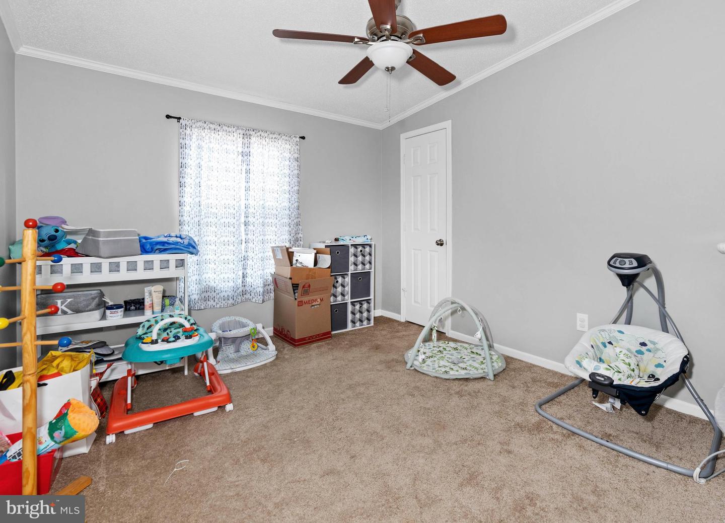 Nursery with baby play equipment, toy storage, patterned curtains, and a brown-bladed ceiling fan.