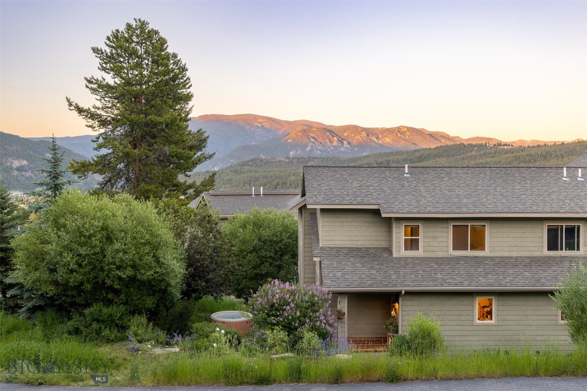 Two-story gray home beneath a mountain range at golden hour, framed by a tall pine and garden greenery.