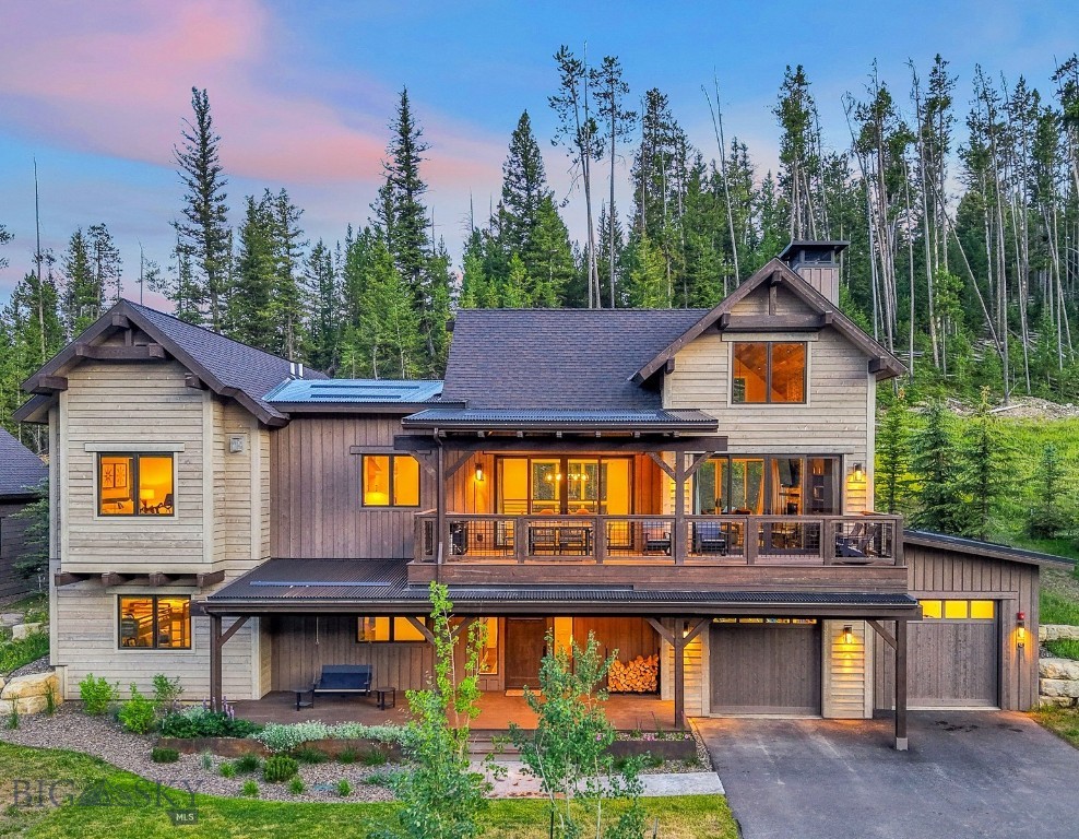 Two-story mountain home with a covered deck, glowing windows, and attached garages at dusk.