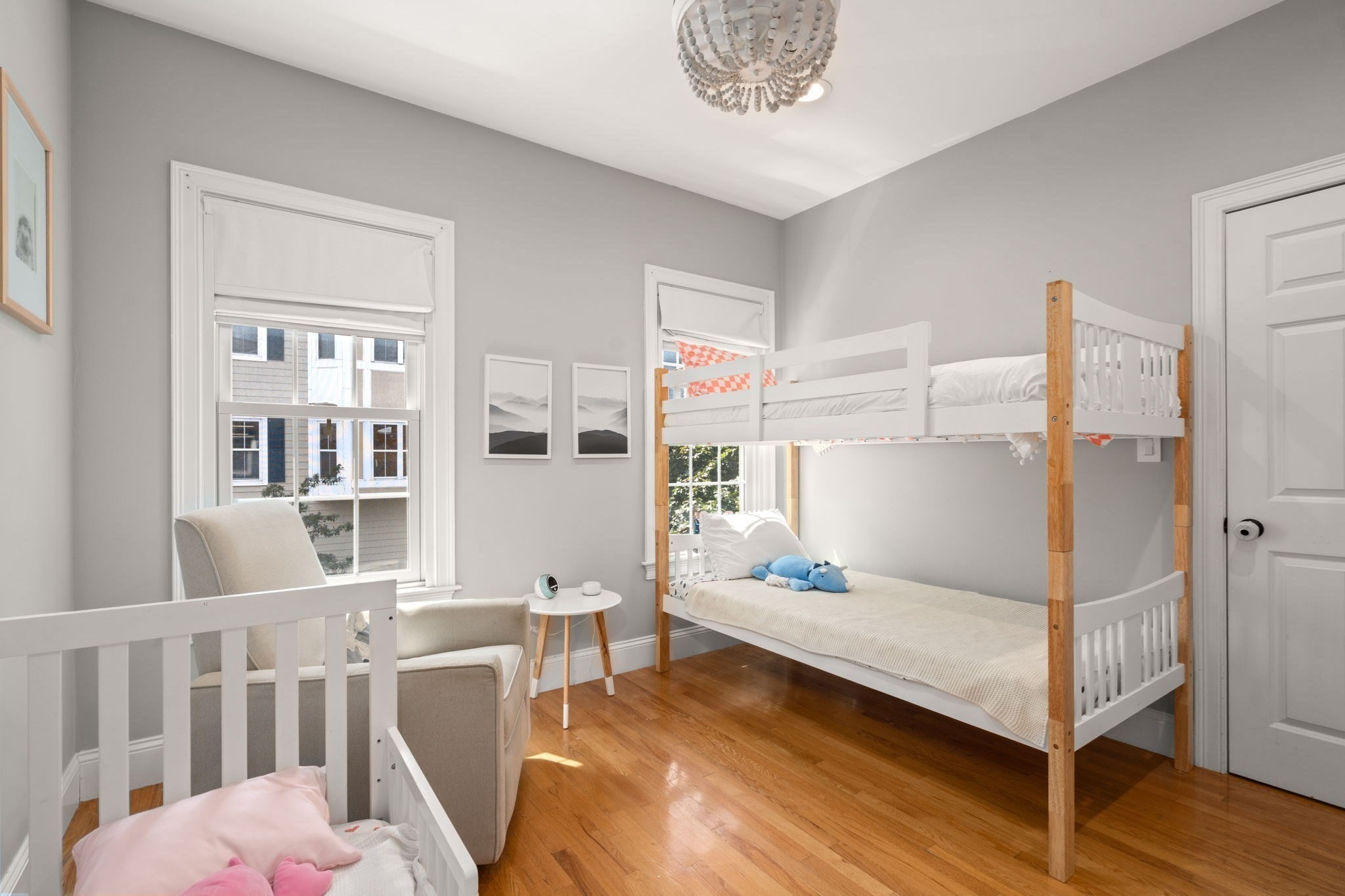 Bedroom with a white-and-wood bunk bed, crib, armchair, and warm hardwood floors.