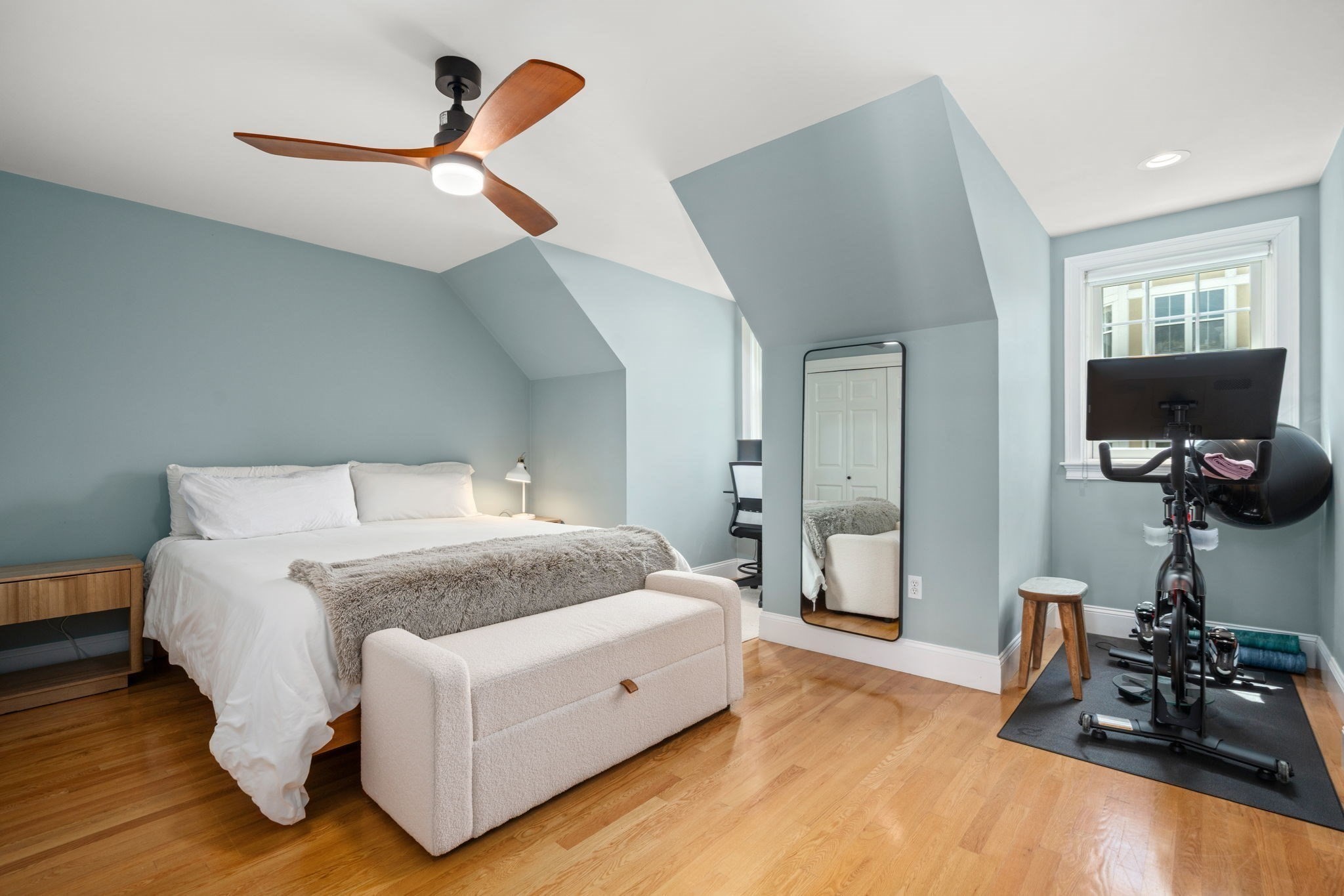 Attic bedroom with a white bed, hardwood floors, and an exercise bike by the window.