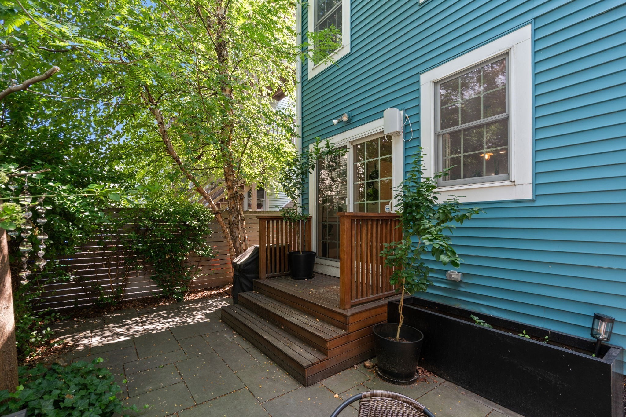 Private patio with a wooden deck, potted plants, and leafy trees beside a blue-sided home.