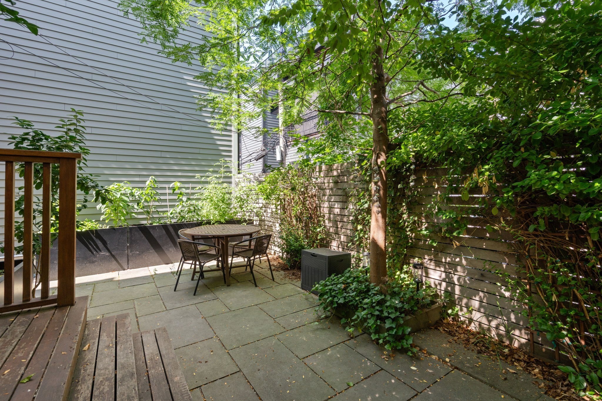 Private paved patio with a round table and chairs, shaded by trees beside a wood privacy fence.