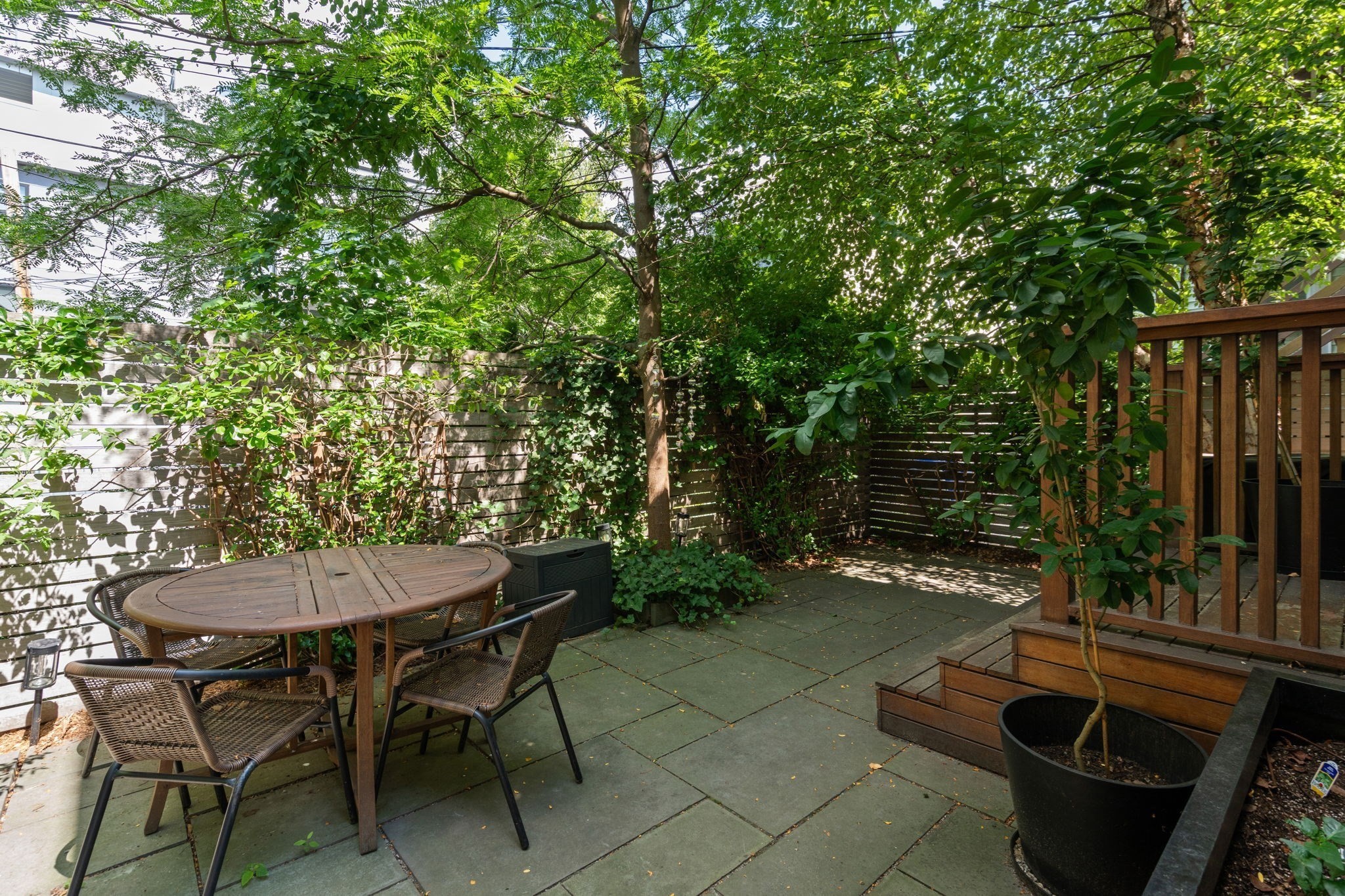 Shaded stone patio with a round dining table, dense greenery, and steps to a wooden deck.