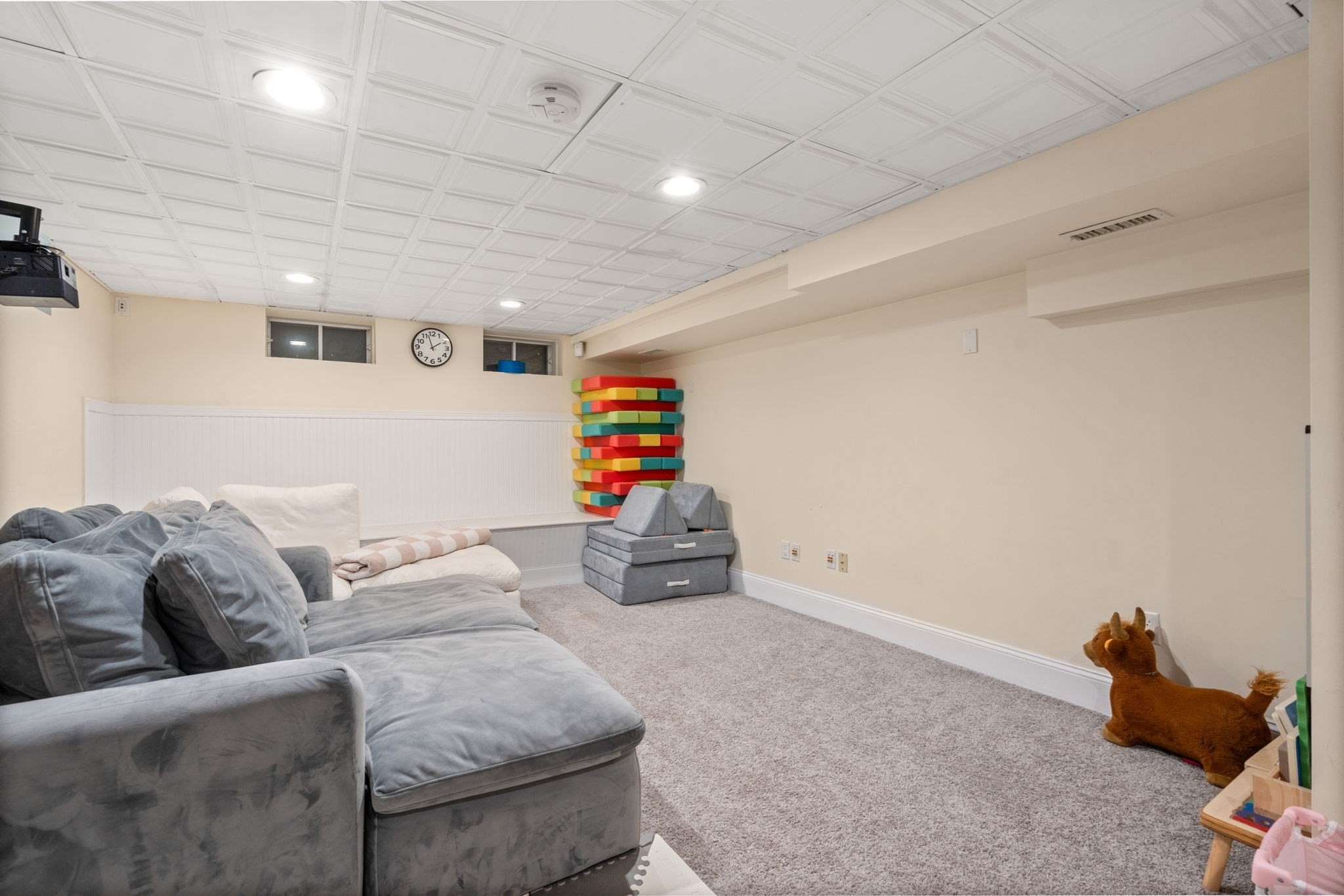 Furnished basement playroom with a gray sectional, carpet, colorful foam blocks, and children’s toys.