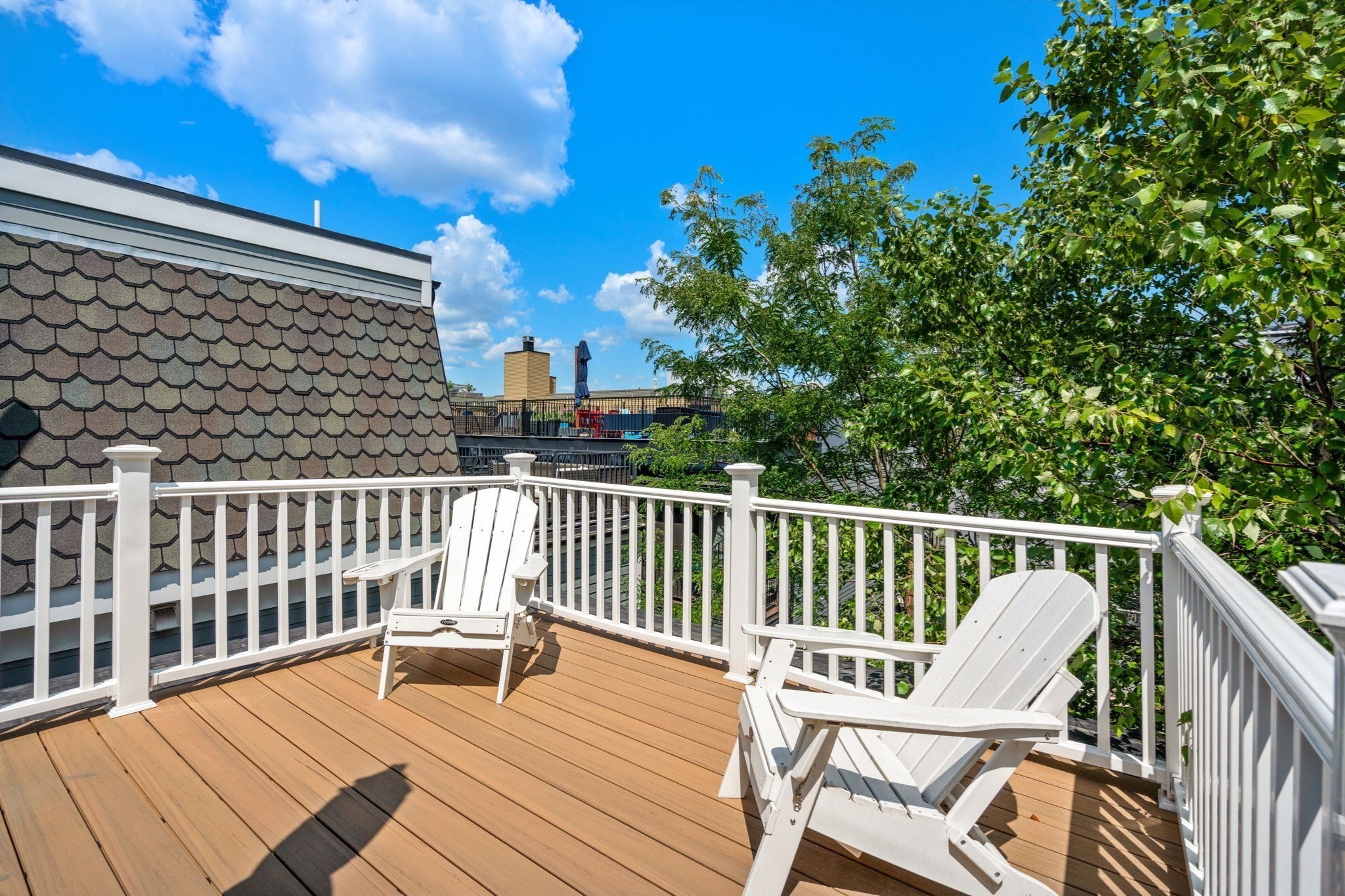 Outdoor deck with tan flooring, white railings, and two Adirondack chairs beside leafy trees.