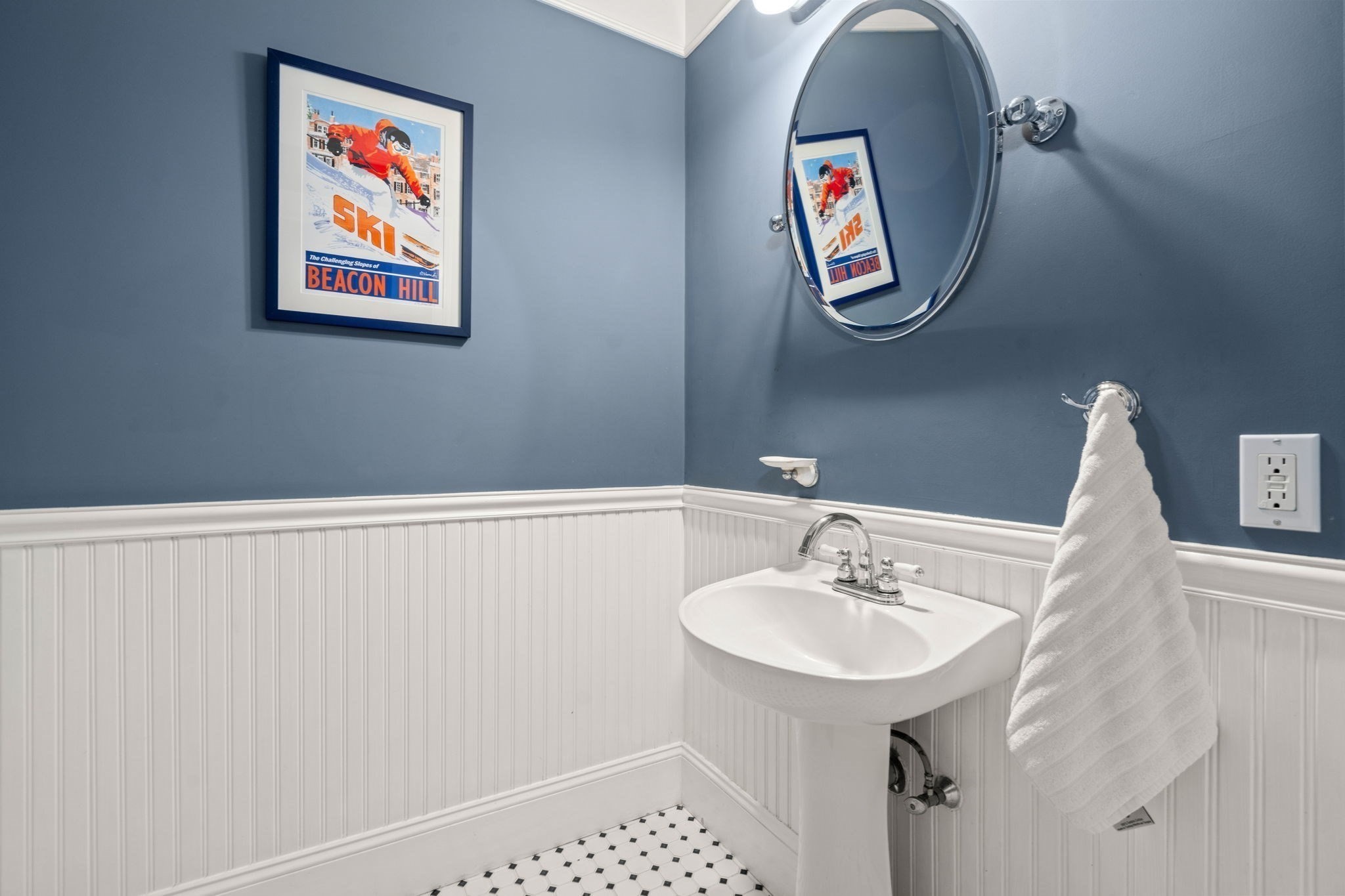 Bathroom with a pedestal sink, oval mirror, white beadboard, and framed Beacon Hill ski poster.