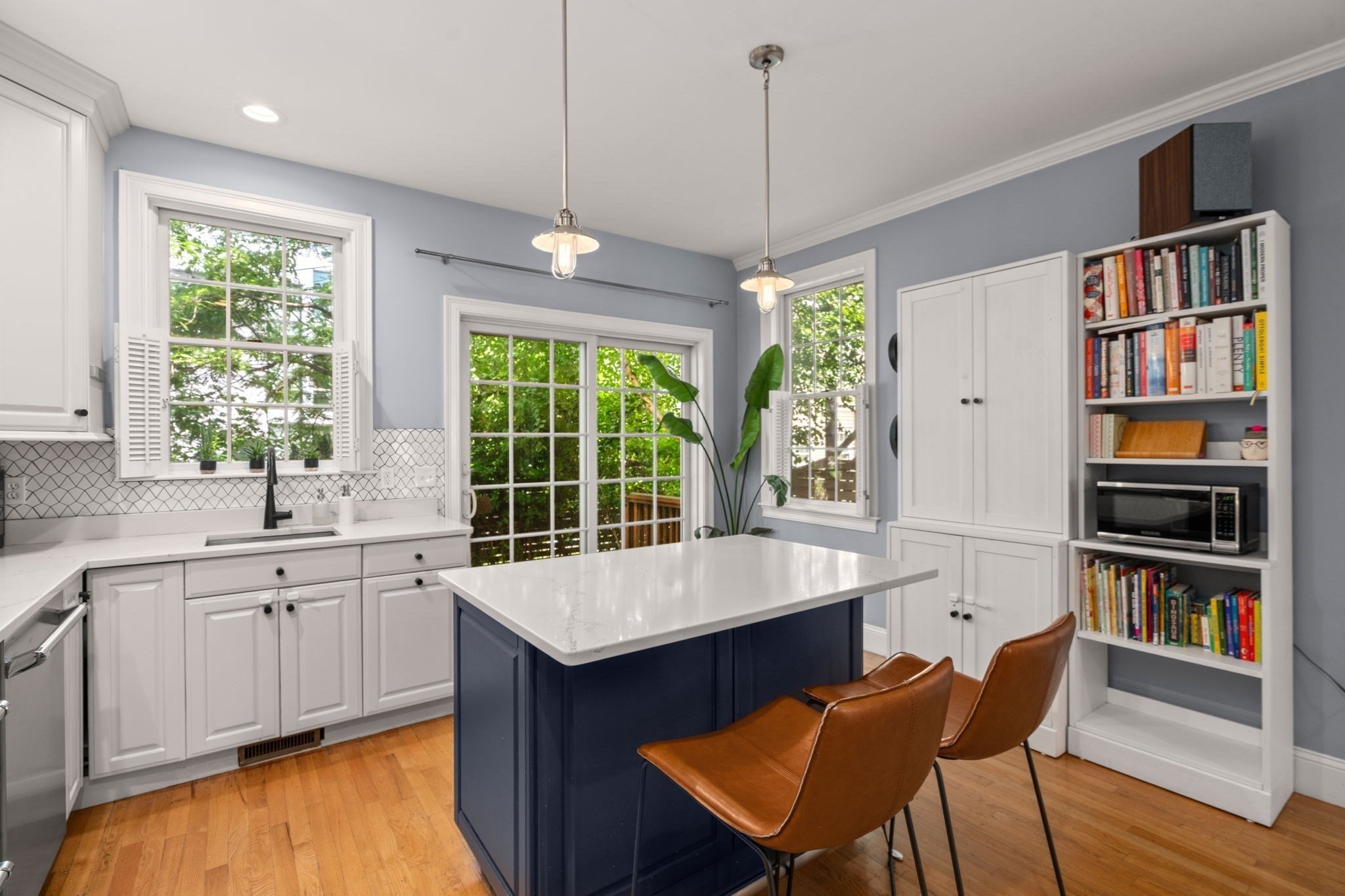 Kitchen with a navy island, brown leather barstools, white cabinetry, and leafy views through large windows.