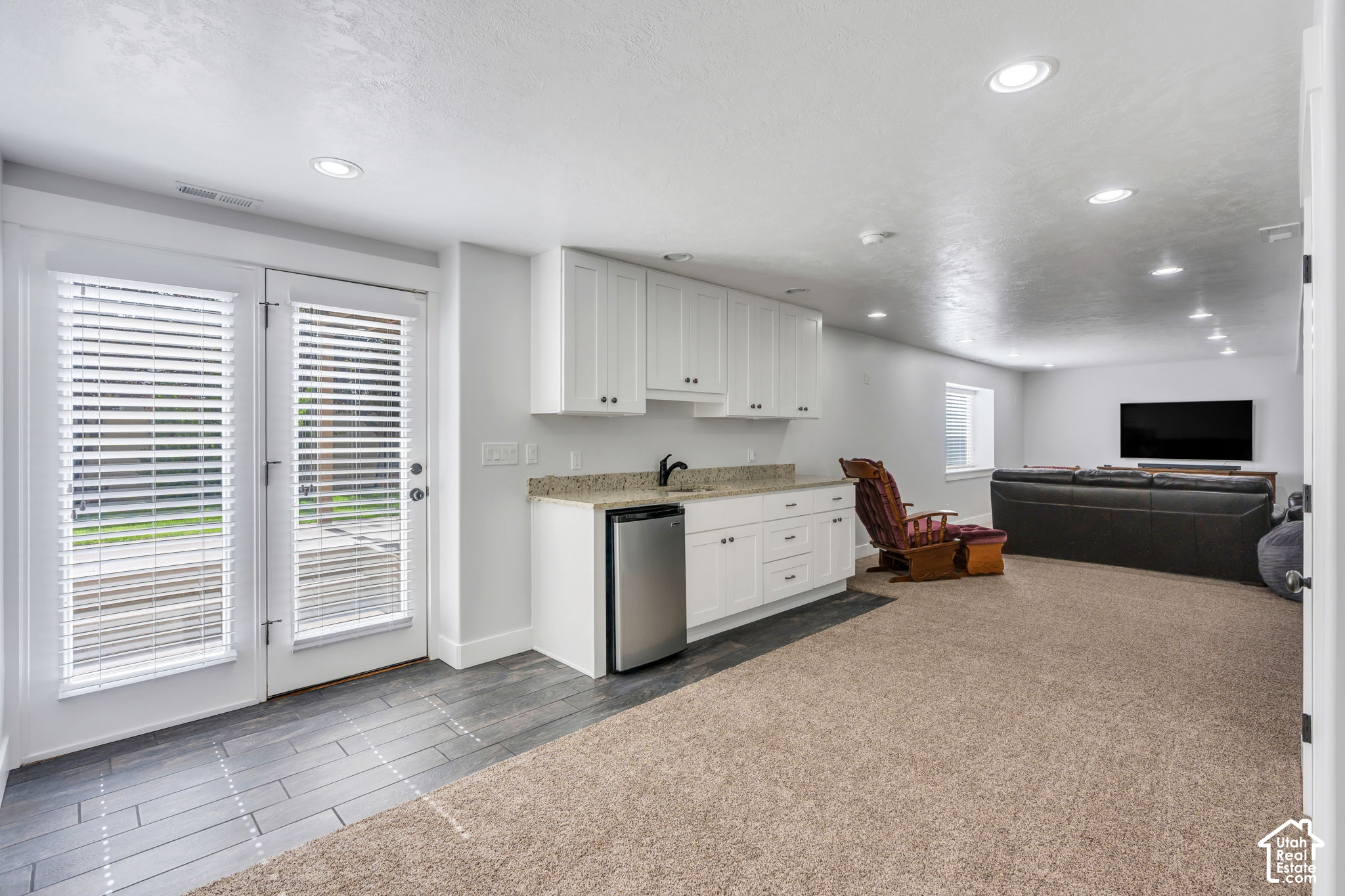 Open living area with a compact white kitchenette, carpeted lounge, black sofa, and wooden recliner.