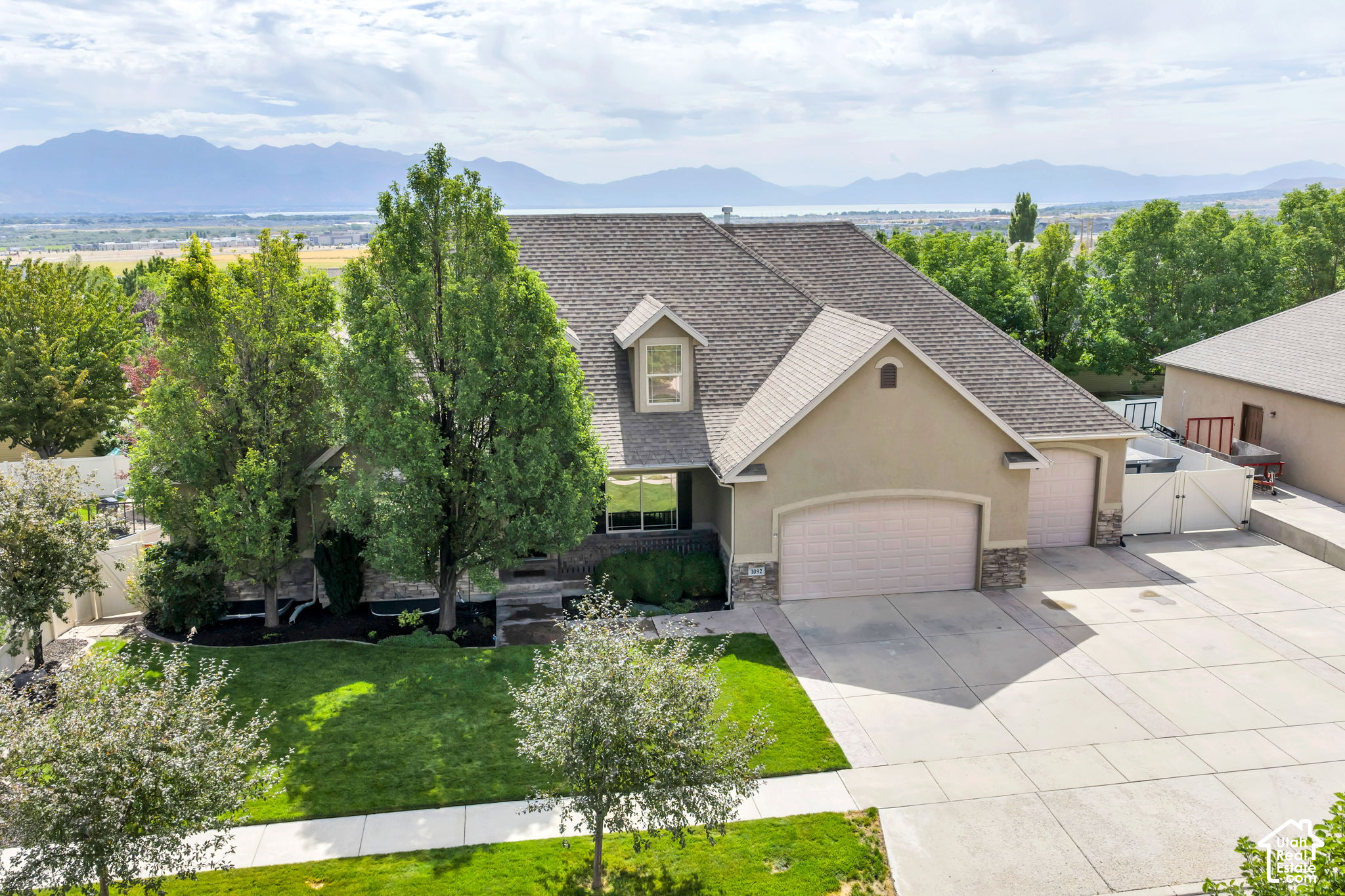Aerial view of a beige home with a broad driveway, leafy landscaping, and distant mountain views.