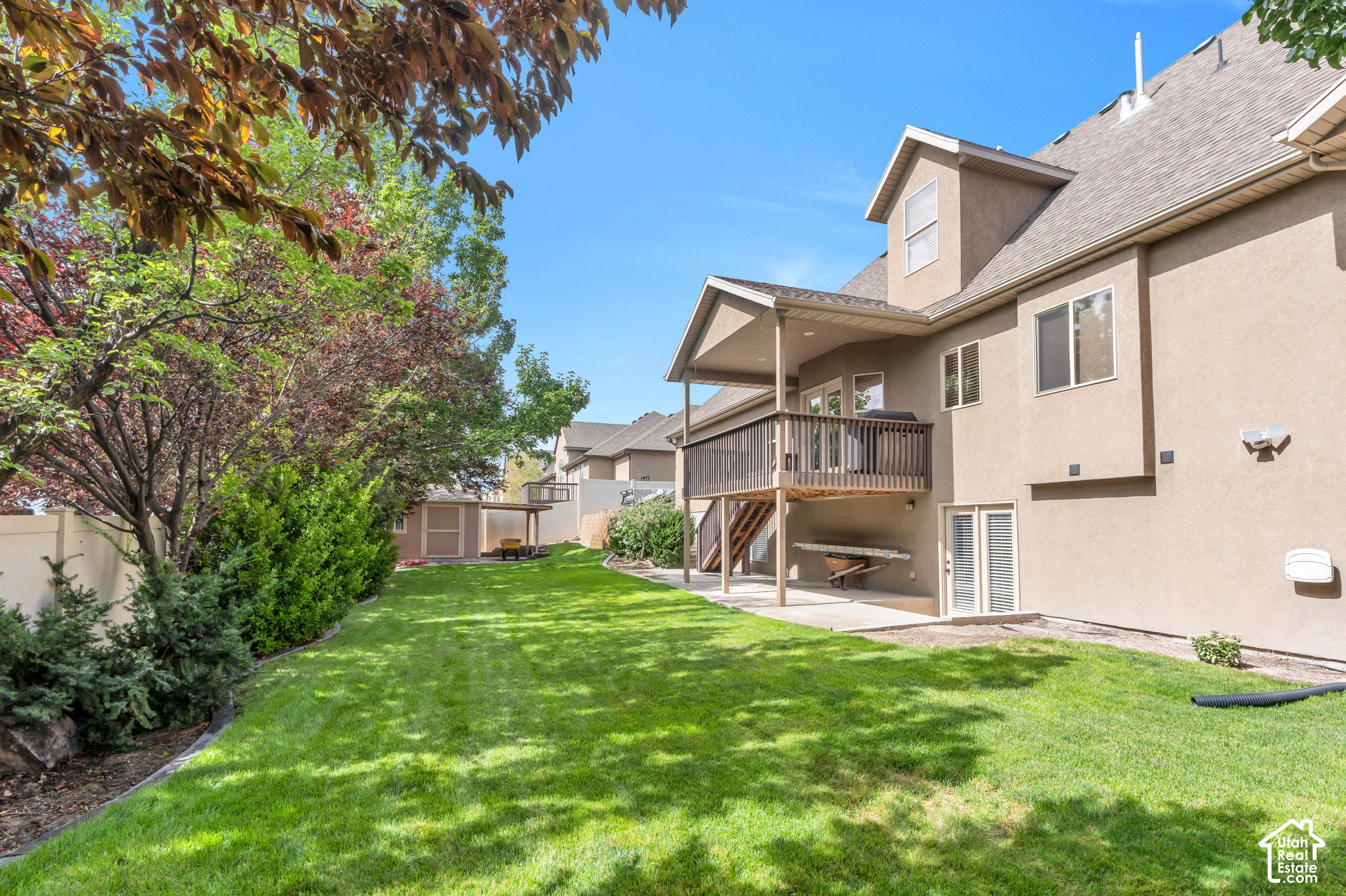 Home’s backyard with a raised deck, broad lawn, mature trees, and a small shed.