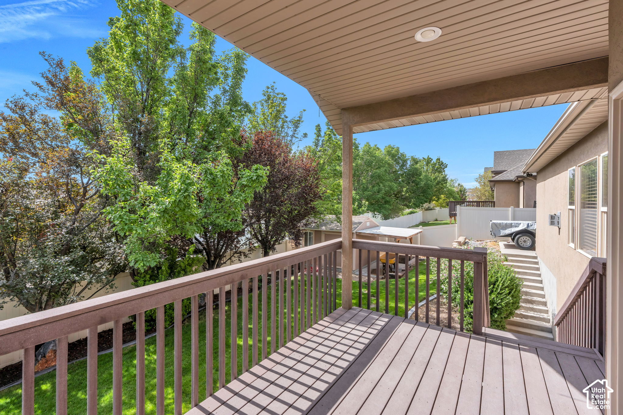 Covered brown deck overlooking a leafy fenced backyard, with stairs beside the house.