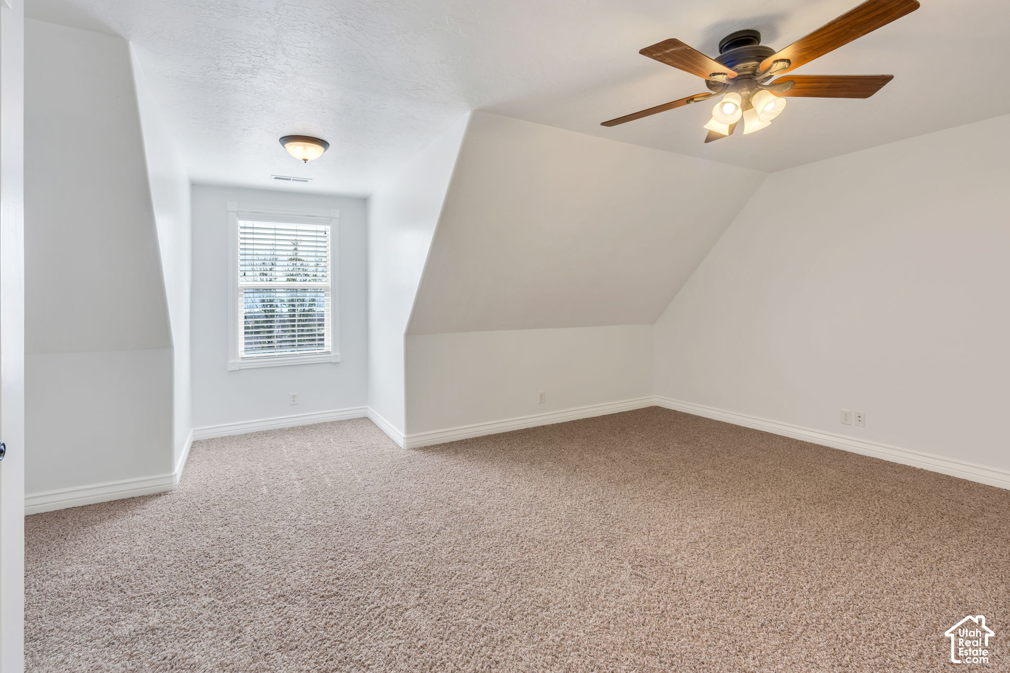 Empty upstairs room with sloped ceilings, beige carpet, a ceiling fan, and a window with white blinds.