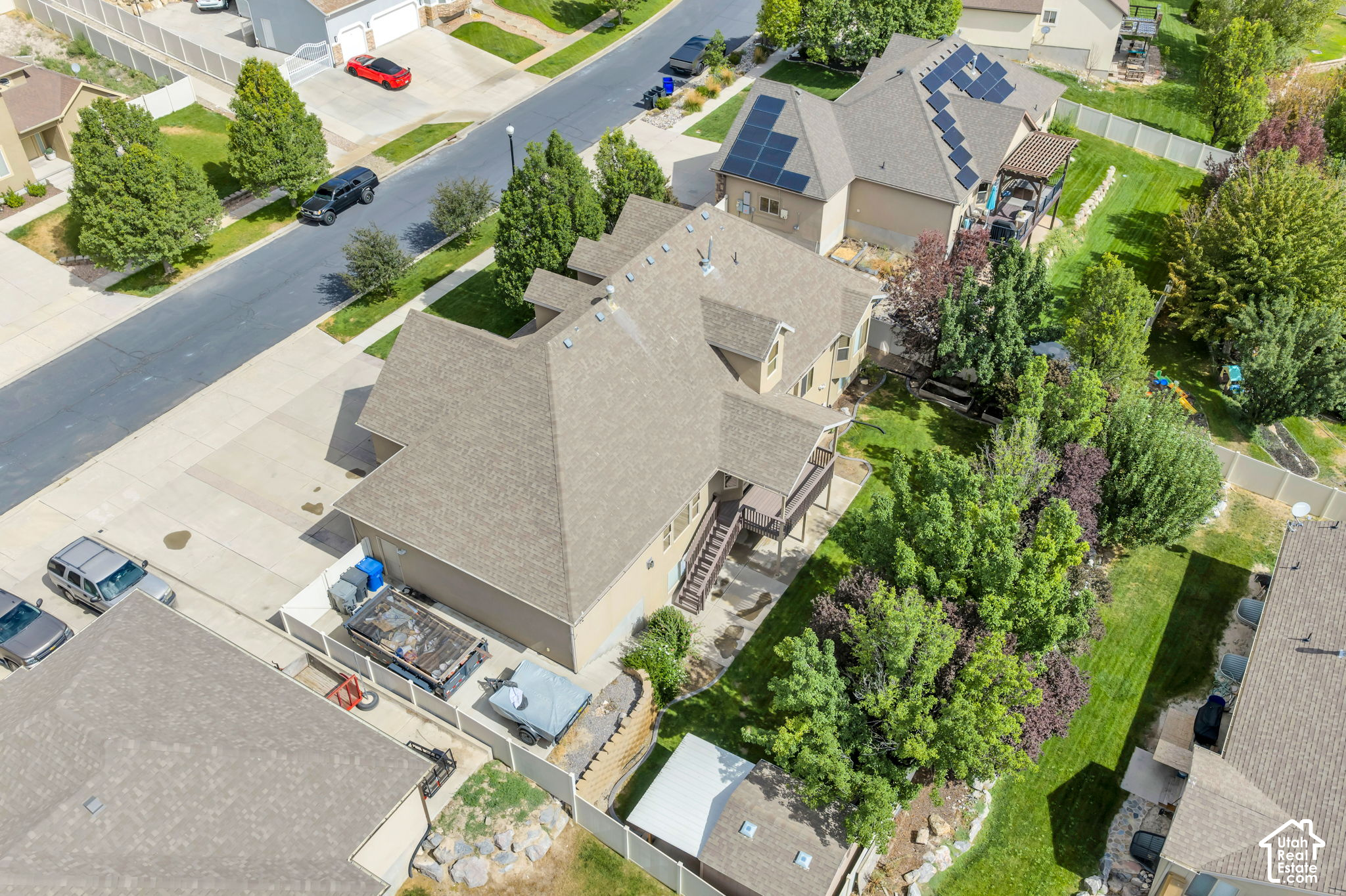 Aerial view of a tan multi-level home with a rear deck, driveway, and mature trees in its backyard.