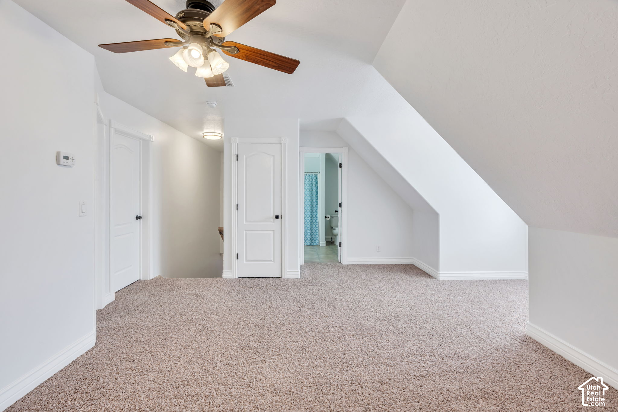 Vacant upstairs bedroom with a sloped ceiling, carpet, ceiling fan, and bathroom doorway.