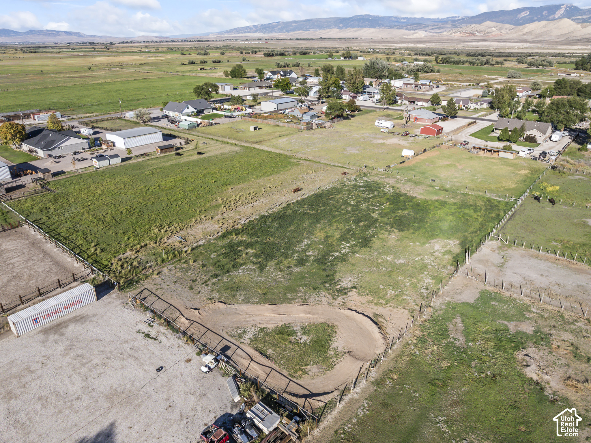 Fenced rural acreage with pasture, a dirt track, nearby homes, and mountains in the distance.