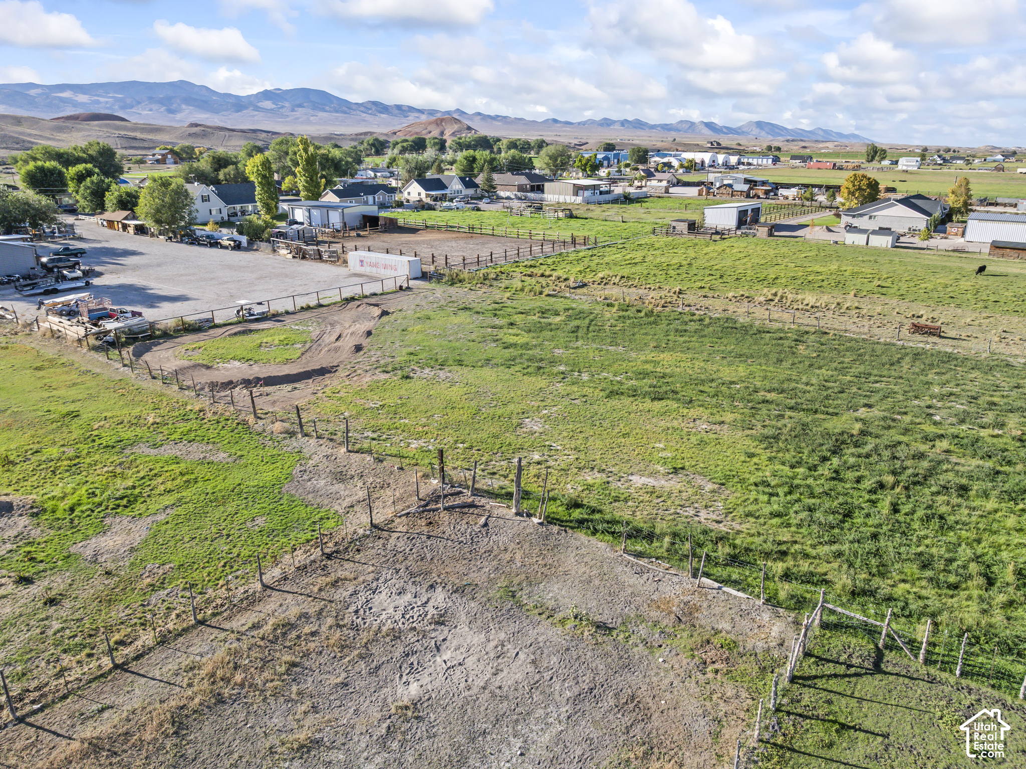 Aerial view of fenced rural land with grassy paddocks, nearby farm buildings, and distant mountains.