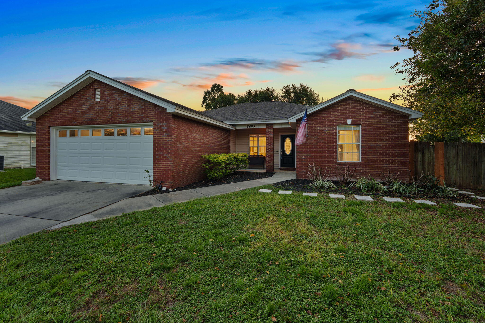 Red brick single-story house with a two-car garage, covered entry, and front lawn at dusk.