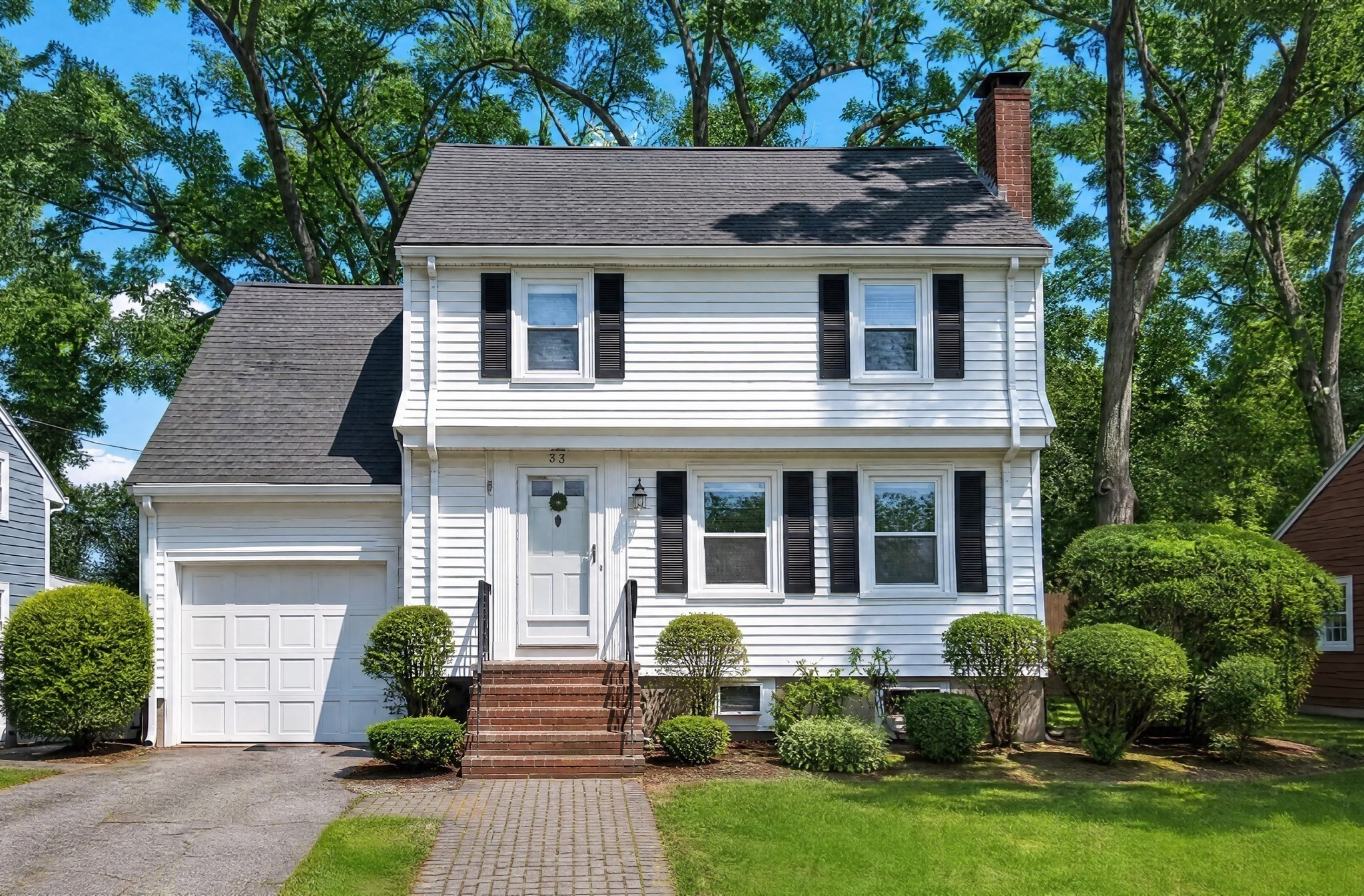 White two-story house with black shutters, attached garage, brick steps, and leafy trees.