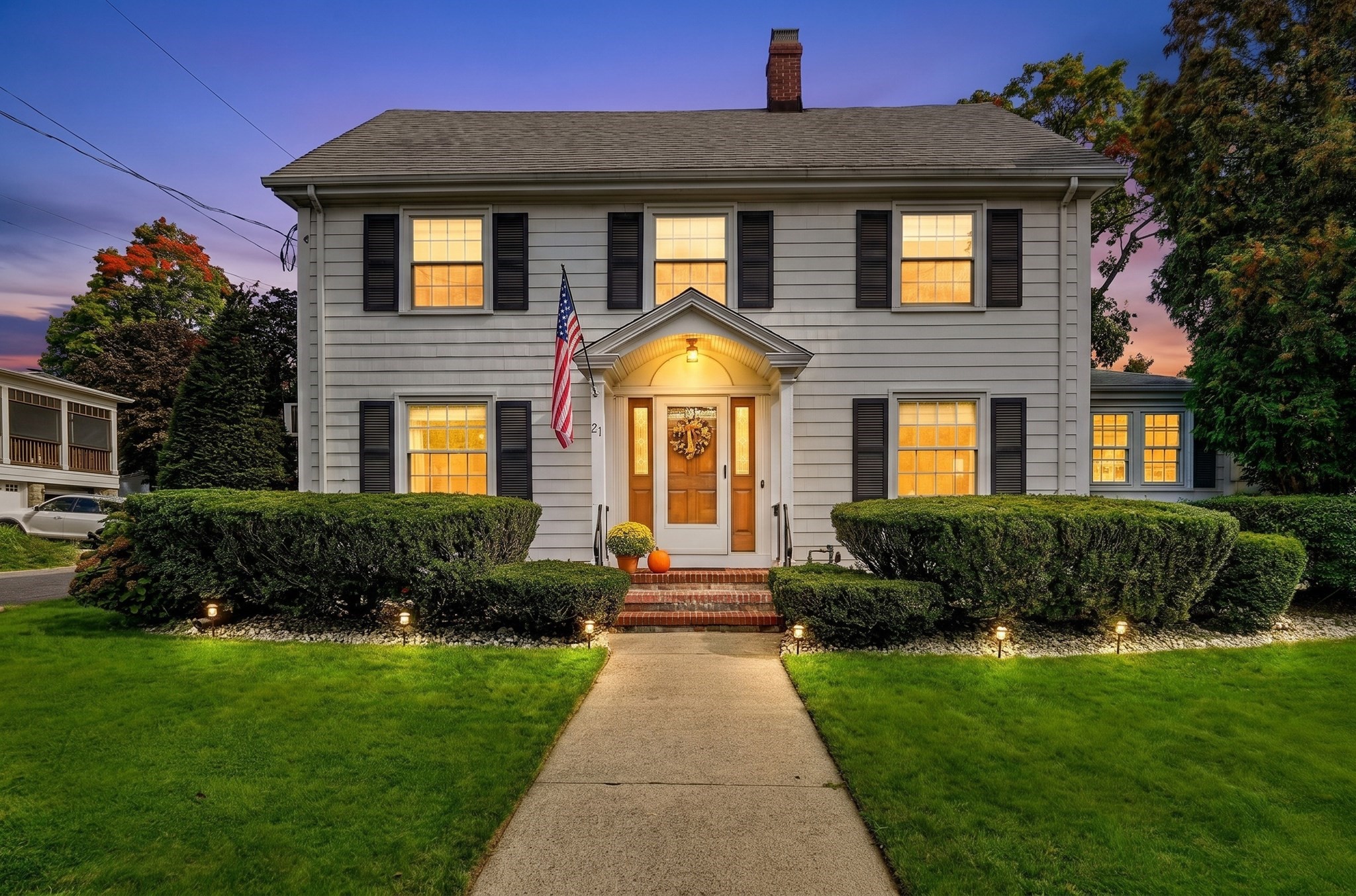 Gray two-story house with glowing windows, black shutters, and autumn decorations at the front entrance.