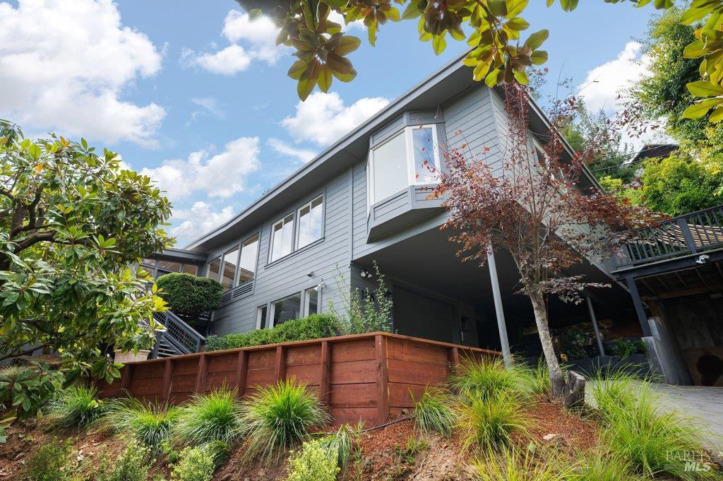 Modern gray two-story home with an elevated deck and landscaped slope behind a redwood retaining wall.