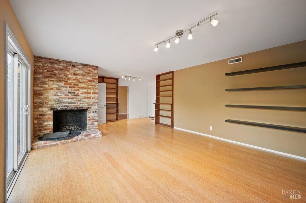 Empty living room with a brick fireplace, built-in shelves, and light wood flooring.