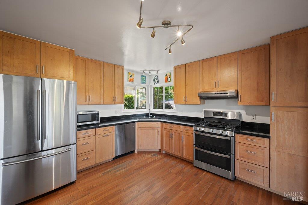 Kitchen with honey-toned cabinets, black countertops, stainless steel appliances, and hardwood floors.