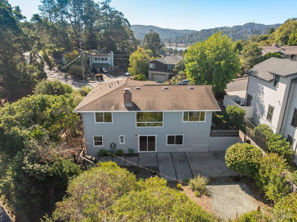 Rear of a gray two-story home with a paved patio, leafy surroundings, and hills in the distance.