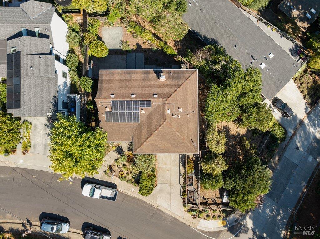 Aerial view of a house with rooftop solar panels, shaded by trees beside a residential street.