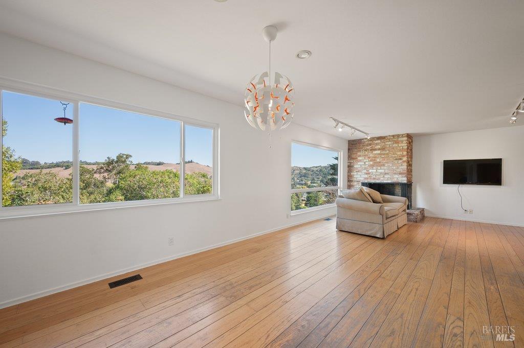 Living room with hardwood floors, large windows overlooking hills, and a brick fireplace.