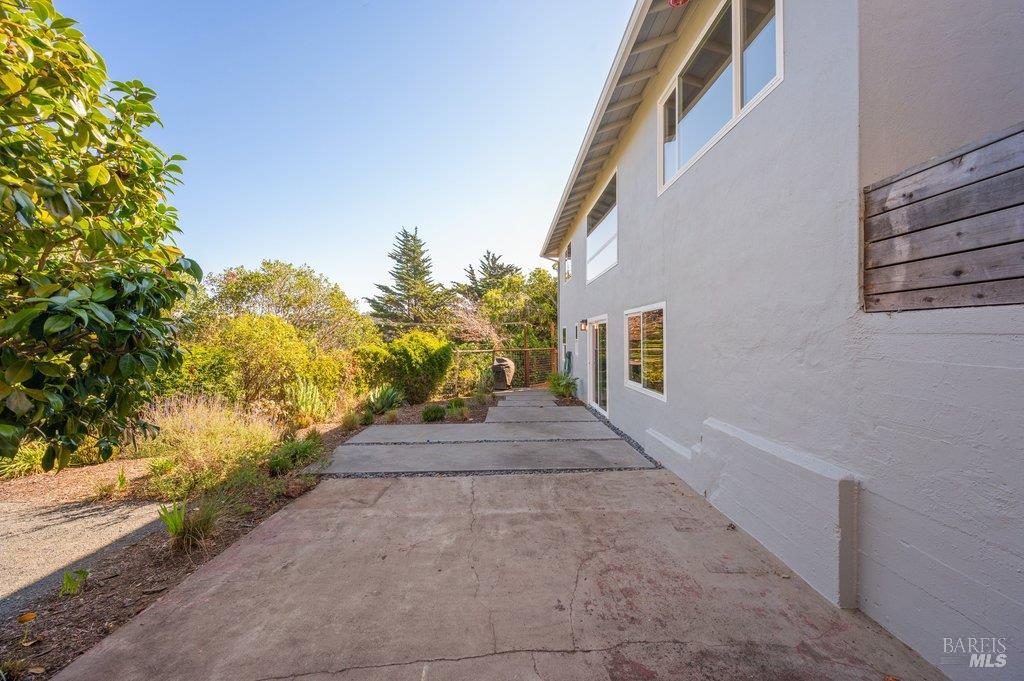 White stucco home beside a broad concrete patio, bordered by shrubs and trees.