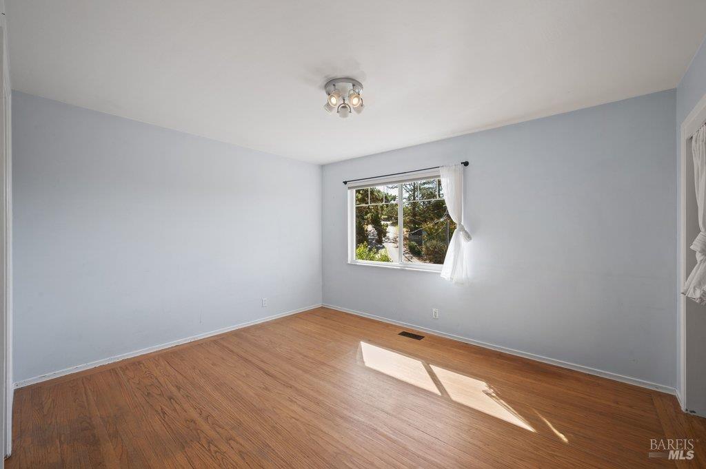 Vacant room with hardwood floors, pale blue walls, and sunlight through a curtained window.