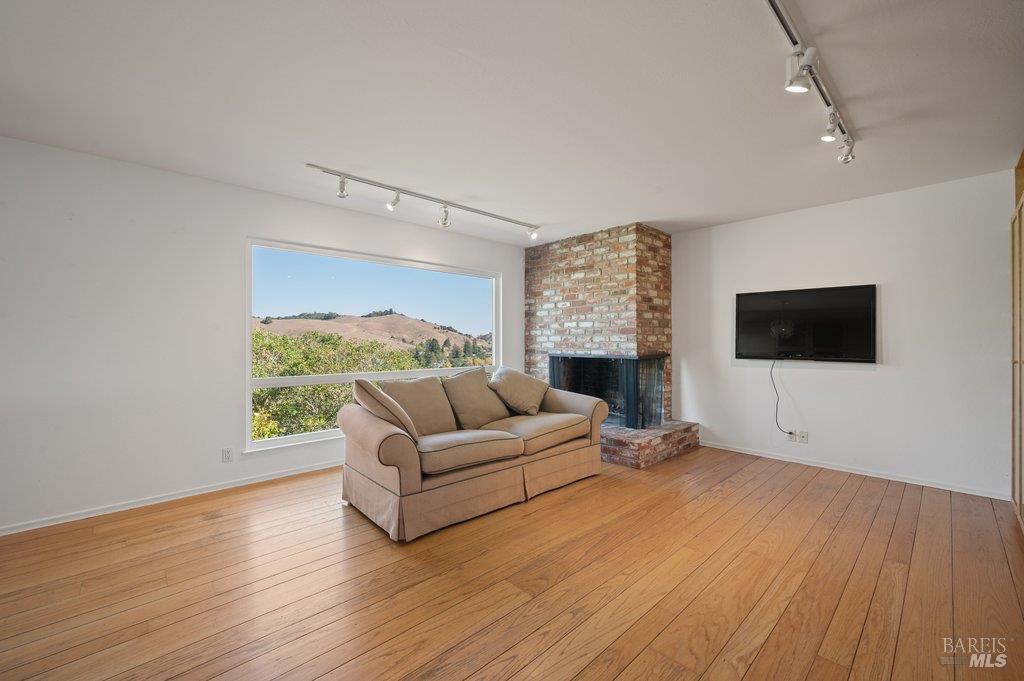 Living room with hardwood floors, brick fireplace, beige sofa, and a large window overlooking rolling hills.