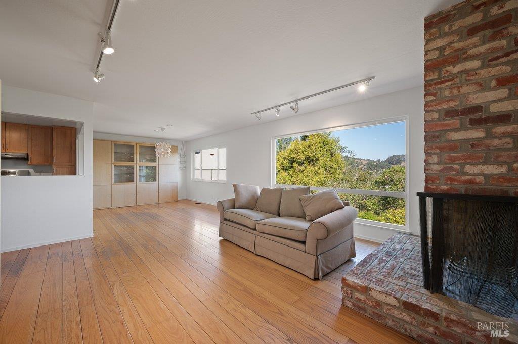 Living room with hardwood floors, brick fireplace, and a large window overlooking trees and hills.