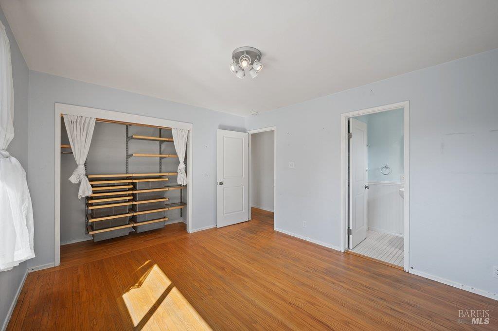 Empty bedroom with hardwood floors, built-in closet shelving, and an open doorway to the bathroom.