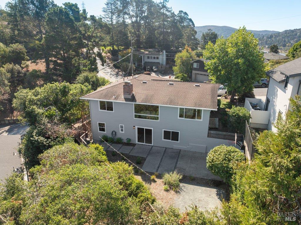 Two-story gray house with a brown roof and driveway, surrounded by trees with hills in the distance.