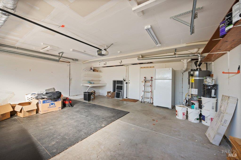 Garage with concrete flooring, overhead door, shelving, refrigerator, and water heater.