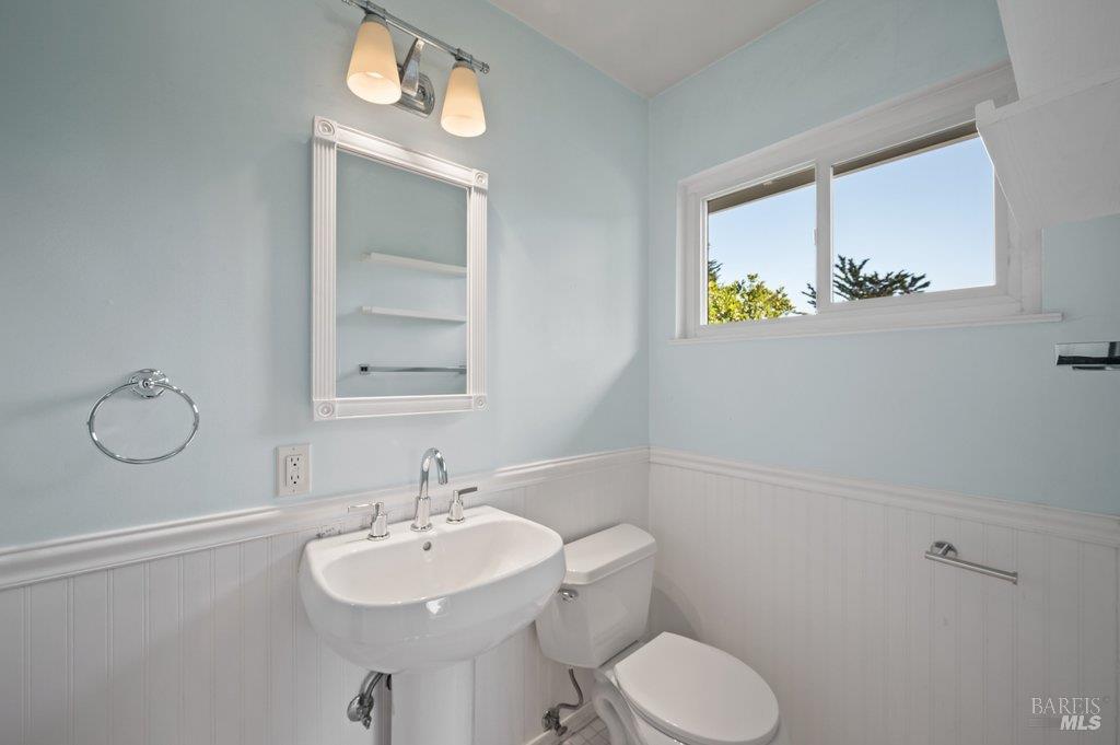 Bathroom with a pedestal sink and toilet, pale blue walls, white beadboard, and a high window.