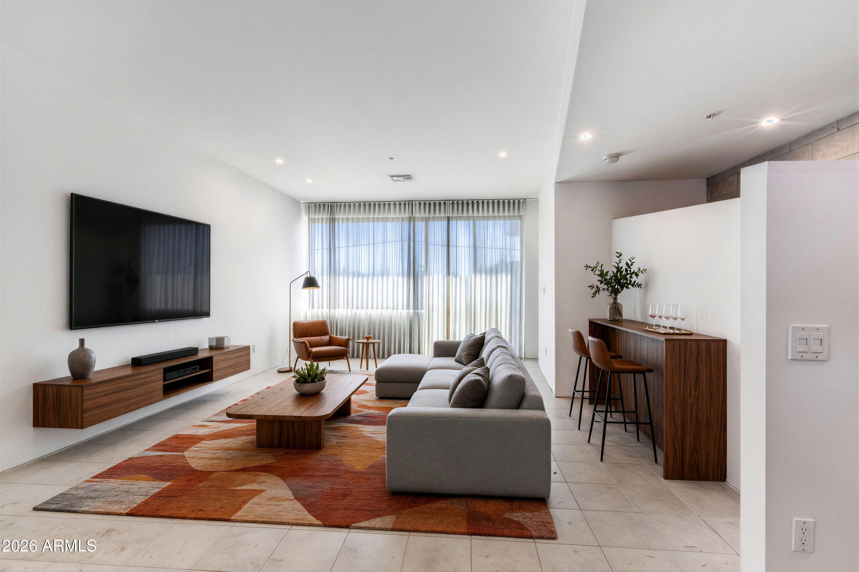 Staged living room with gray sectional, wood furniture, breakfast bar, and wide curtained windows.