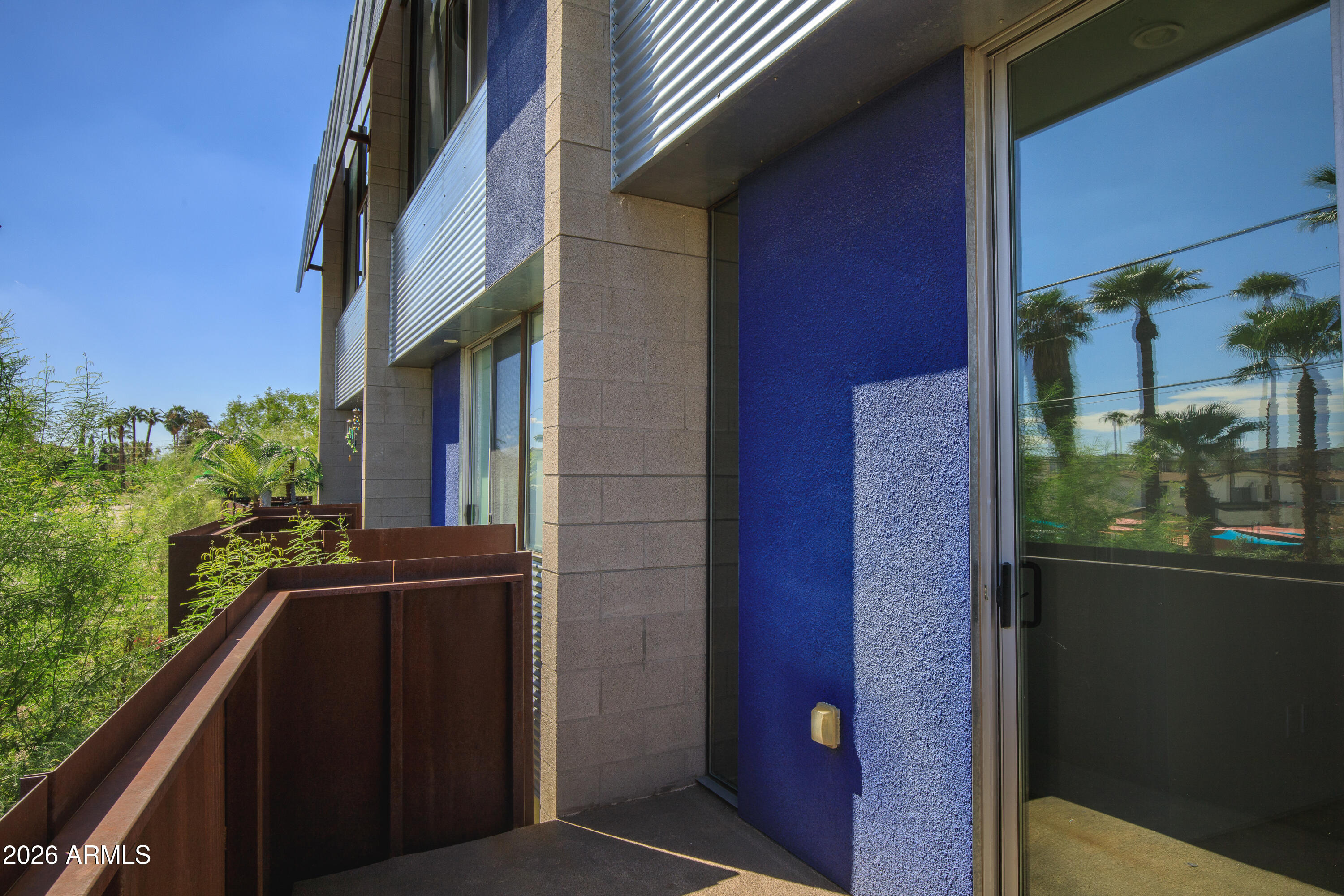 Modern apartment balcony with blue wall panels, rust-colored railing, and palm trees reflected in glass doors.