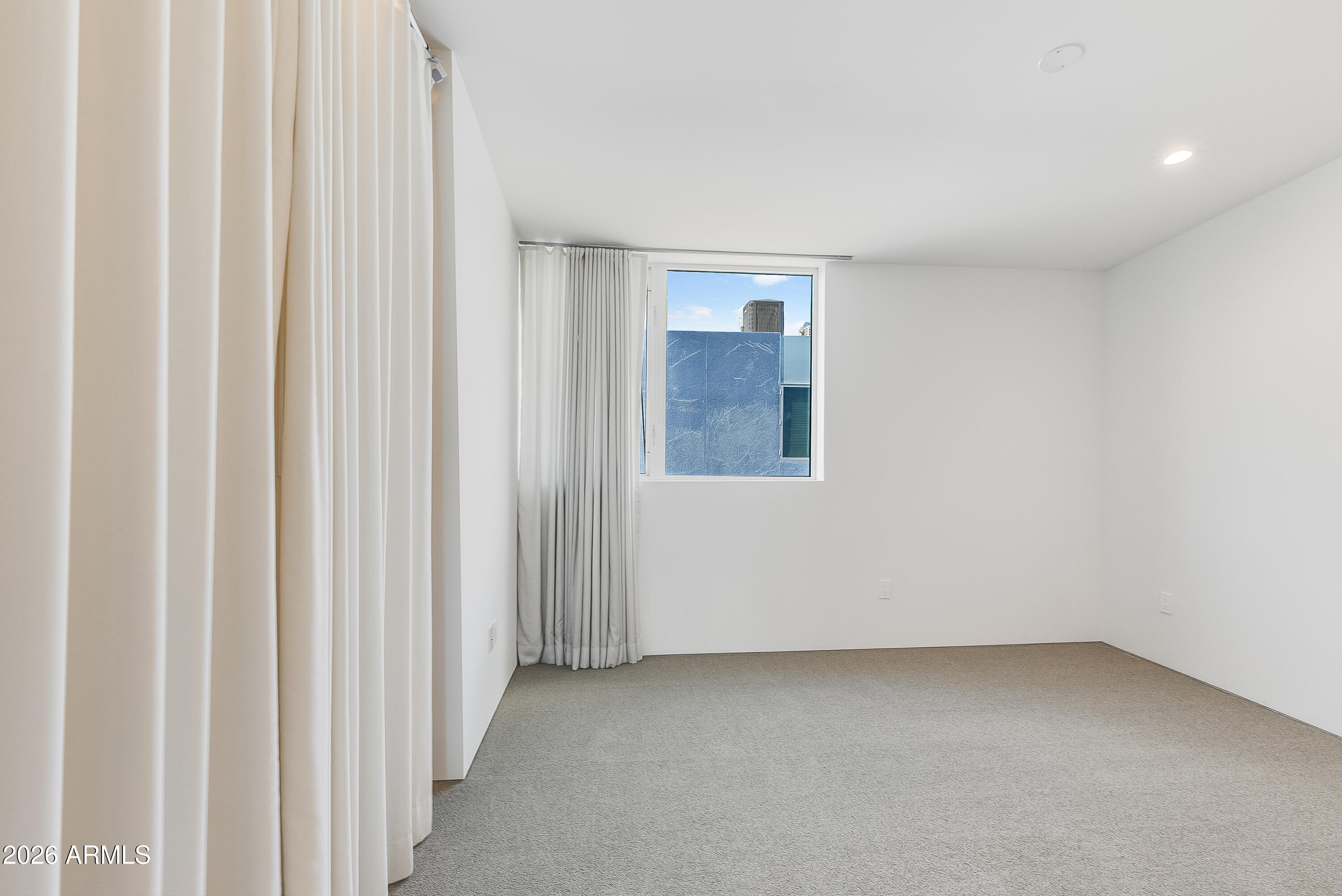 Empty carpeted bedroom with floor-to-ceiling curtains and a window overlooking a nearby building.