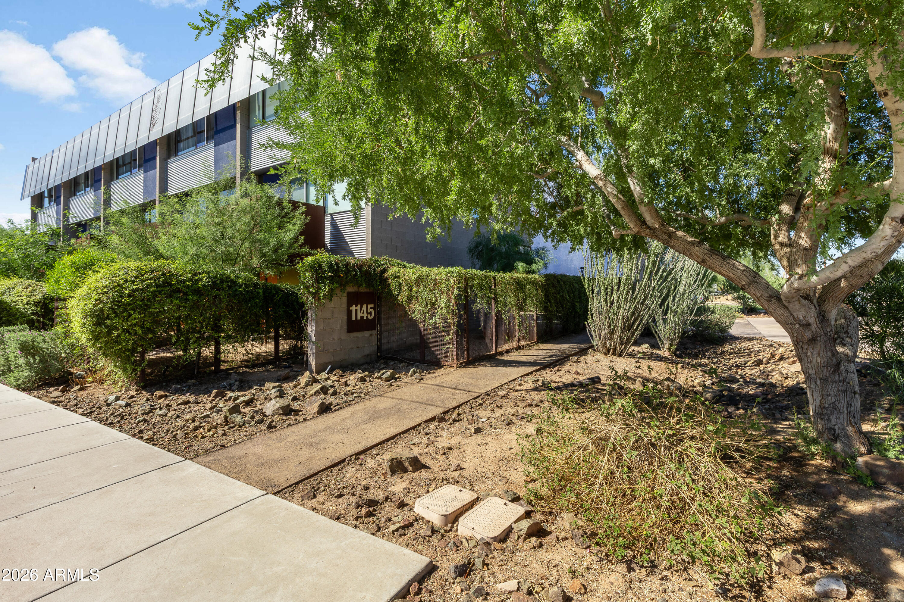 Modern multi-story building behind desert landscaping, with a large shade tree and walkway to the gated entrance.
