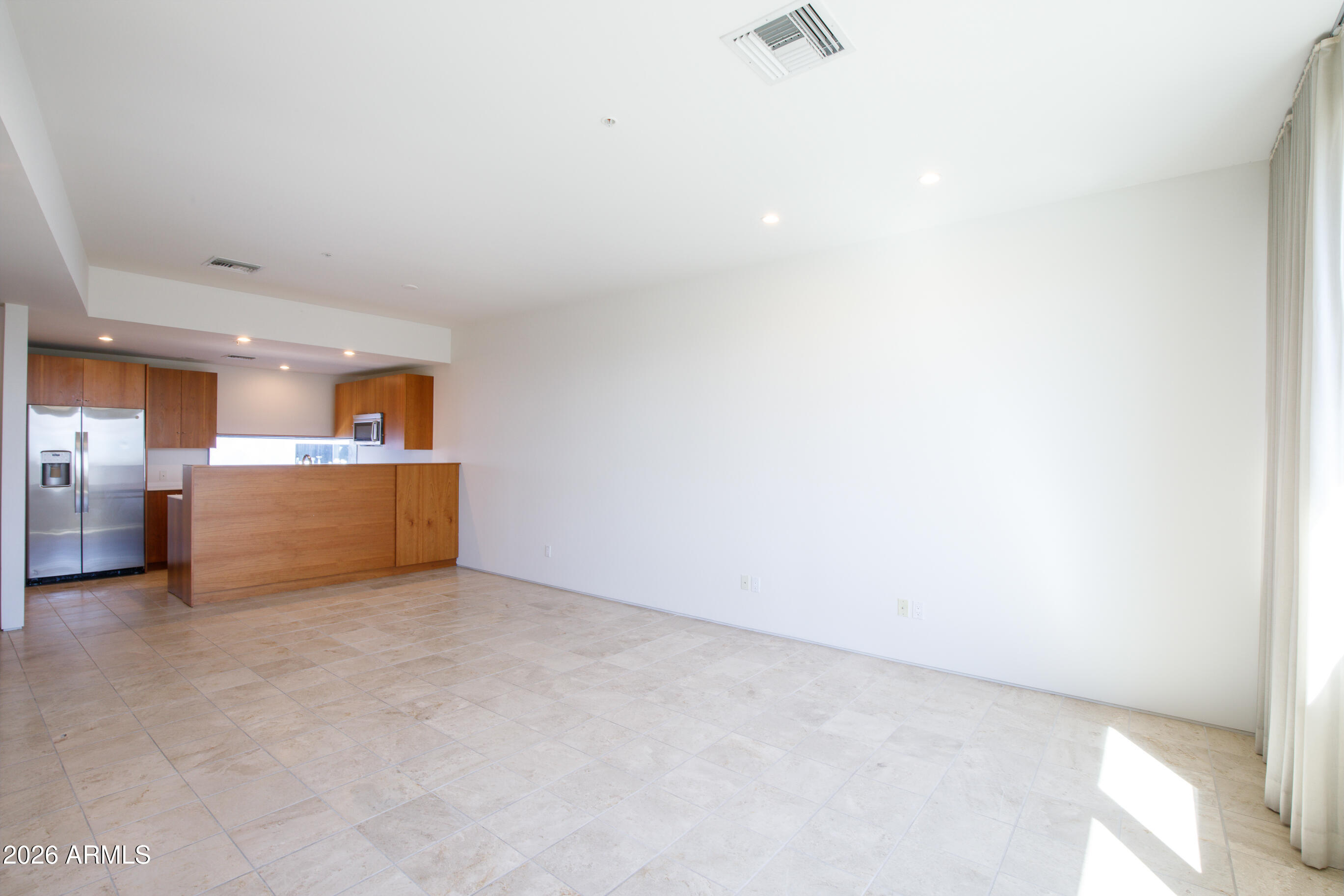 Vacant living area with beige tile floors, wood kitchen peninsula, and stainless steel refrigerator.