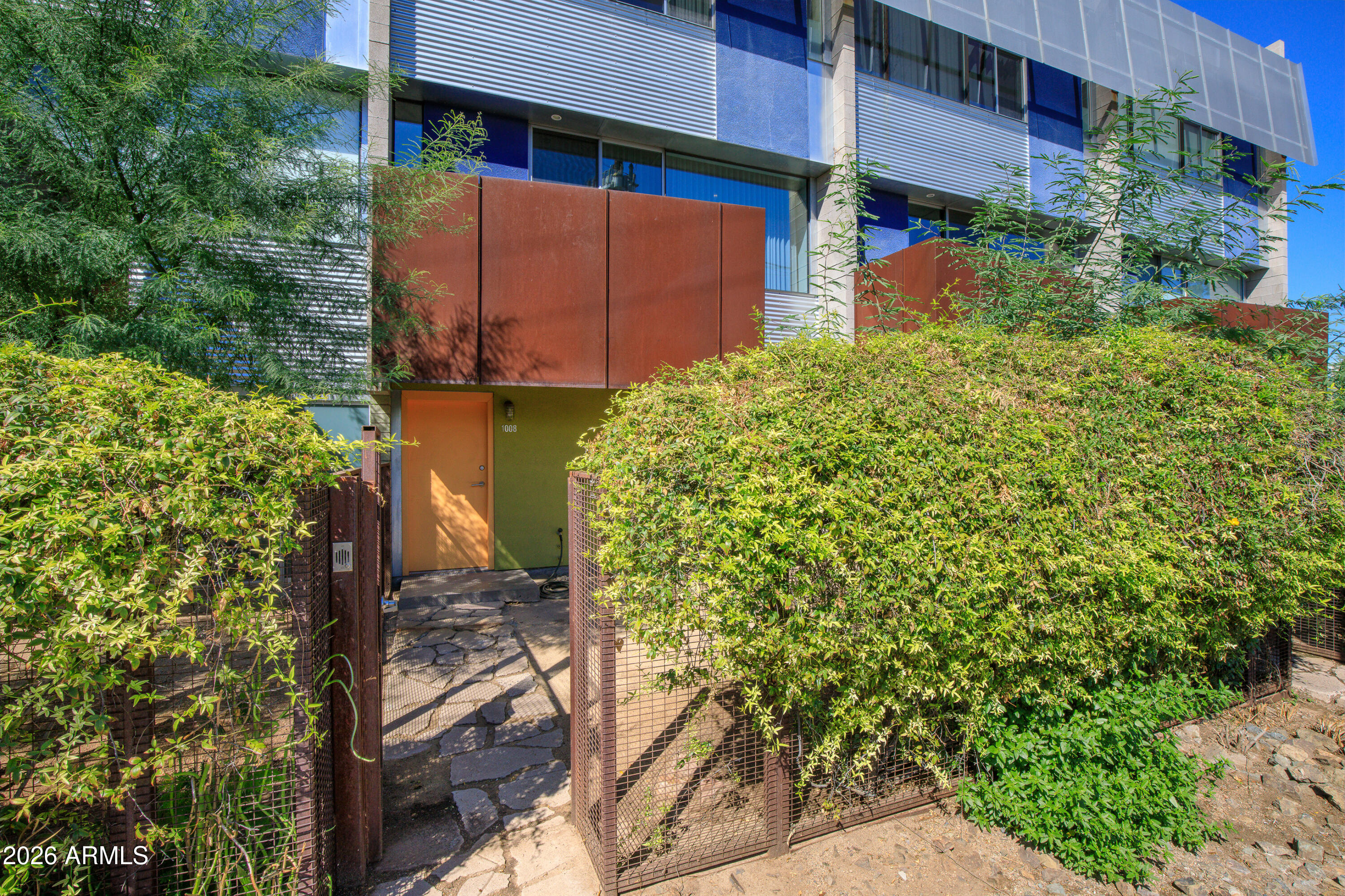 Modern building entrance to unit 1008, framed by dense hedges and rust-colored panels.