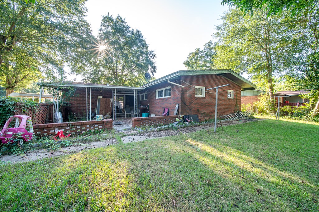 Red brick house with a covered patio and grassy backyard beneath mature trees.
