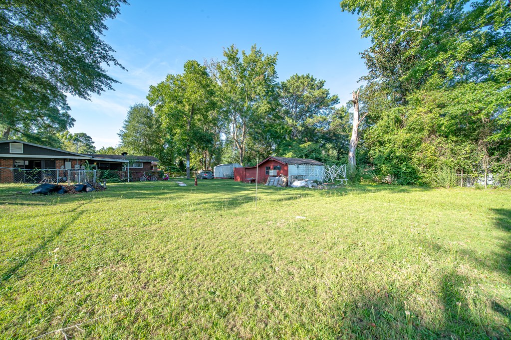 Grassy yard with a single-story brick home, red outbuilding, and mature trees.