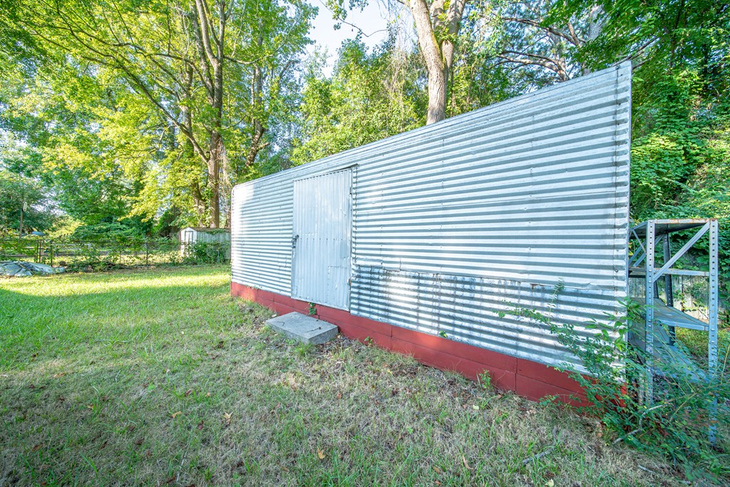 Corrugated-metal shed with a central door and open shelving in a grassy, tree-lined yard.