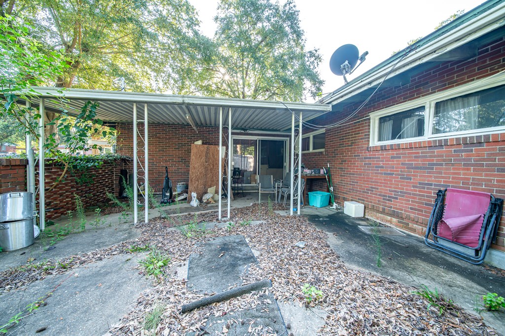 Covered patio beside a red-brick home, with metal roofing and trees along the edge.