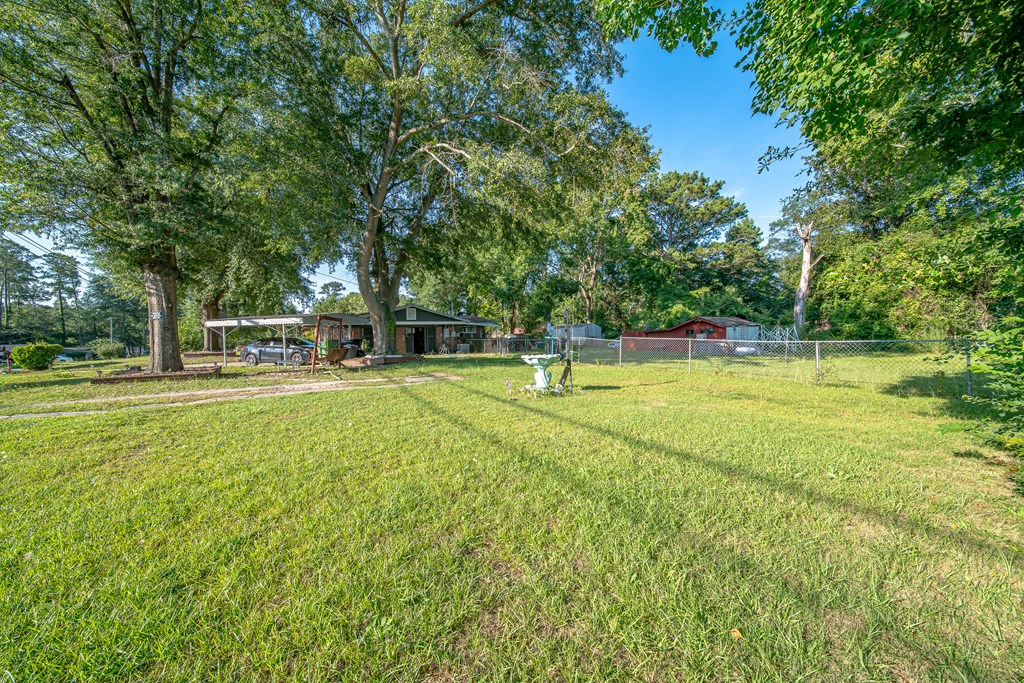 Grassy yard with mature trees, chain-link fencing, and a single-story home with a carport.
