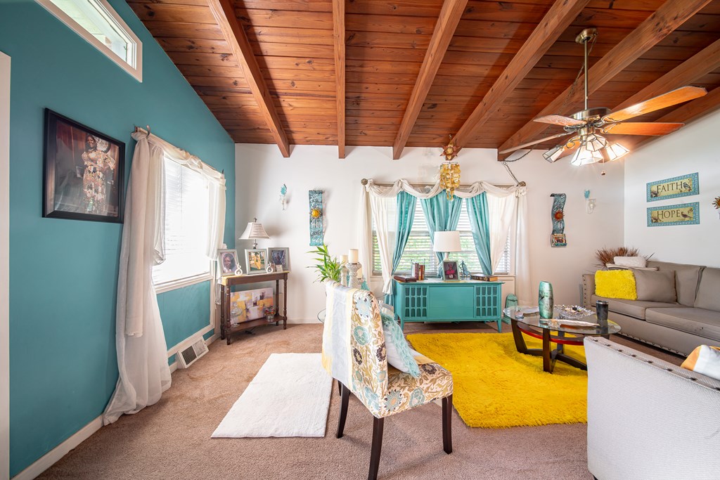 Living room with exposed wood ceiling beams, teal wall, gray sofa, and bright yellow rug.